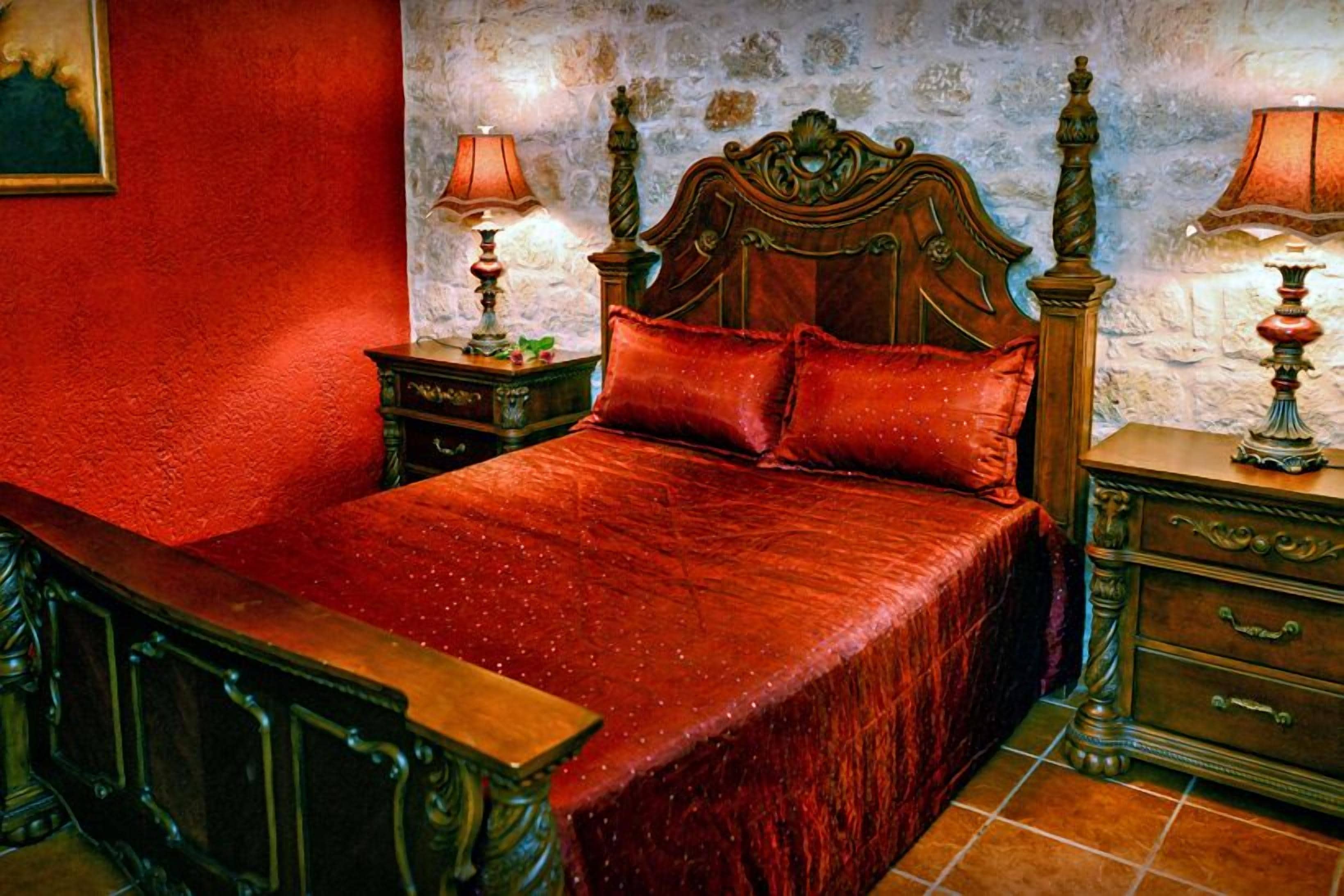 Ein Schlafzimmer mit einem hölzernen Bett, roter Bettwäsche, zwei Nachttischen mit Lampen und einer Steinwand.