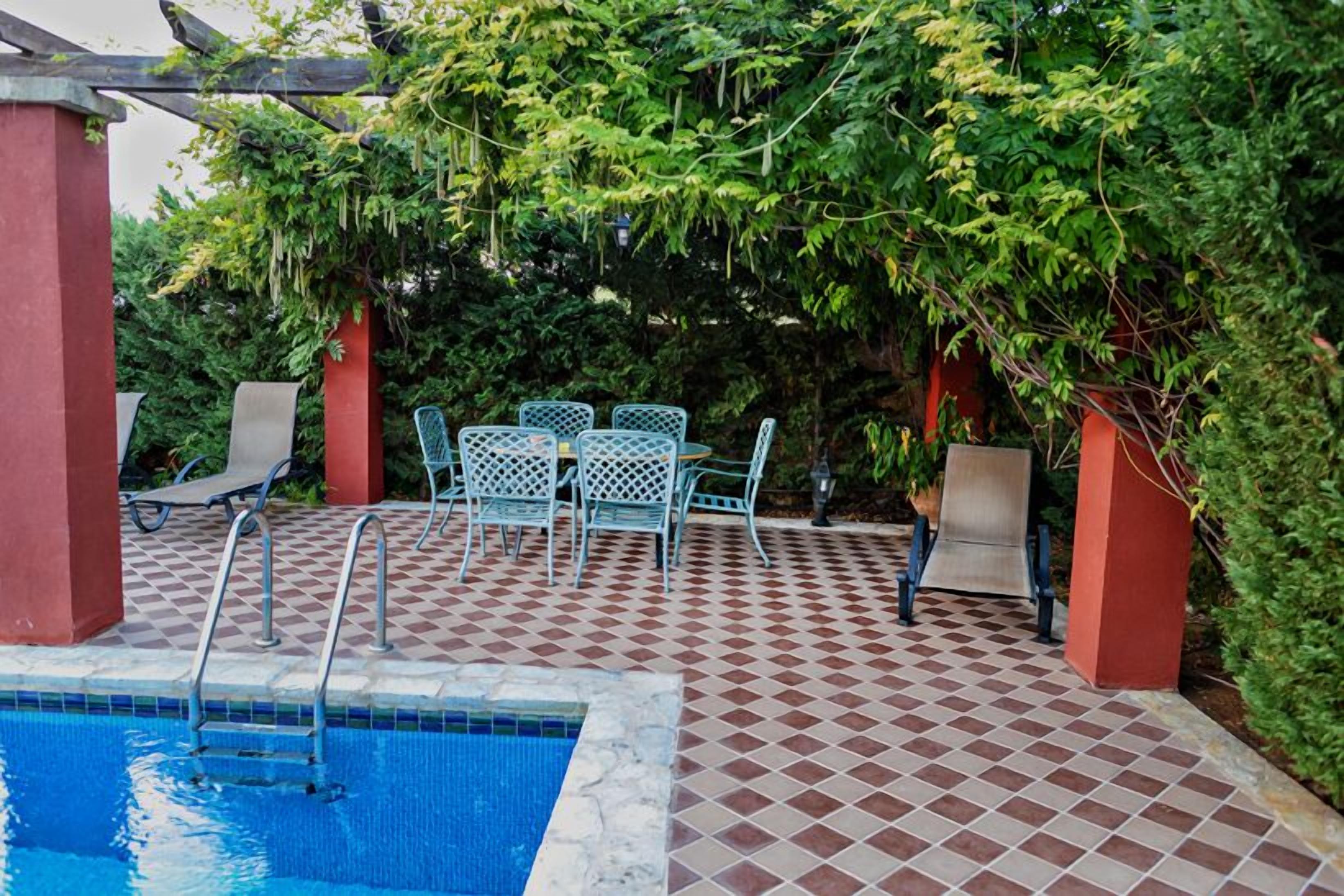 A pool area with a checkered tile floor, metal chairs, and a pergola with greenery.
