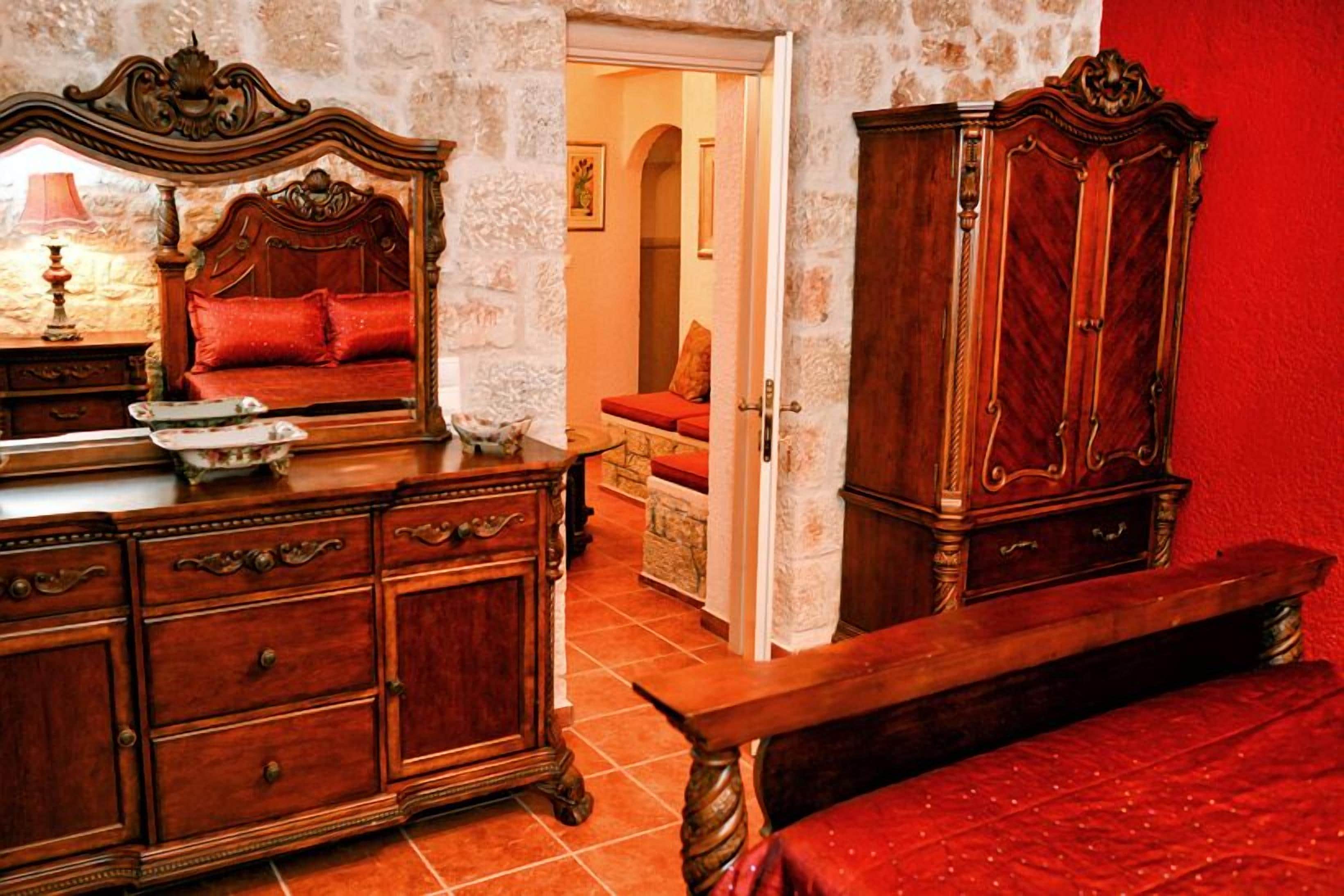 A room with a wooden bed, a wooden dresser, and a wooden wardrobe. There is a red upholstered bench in the foreground.
