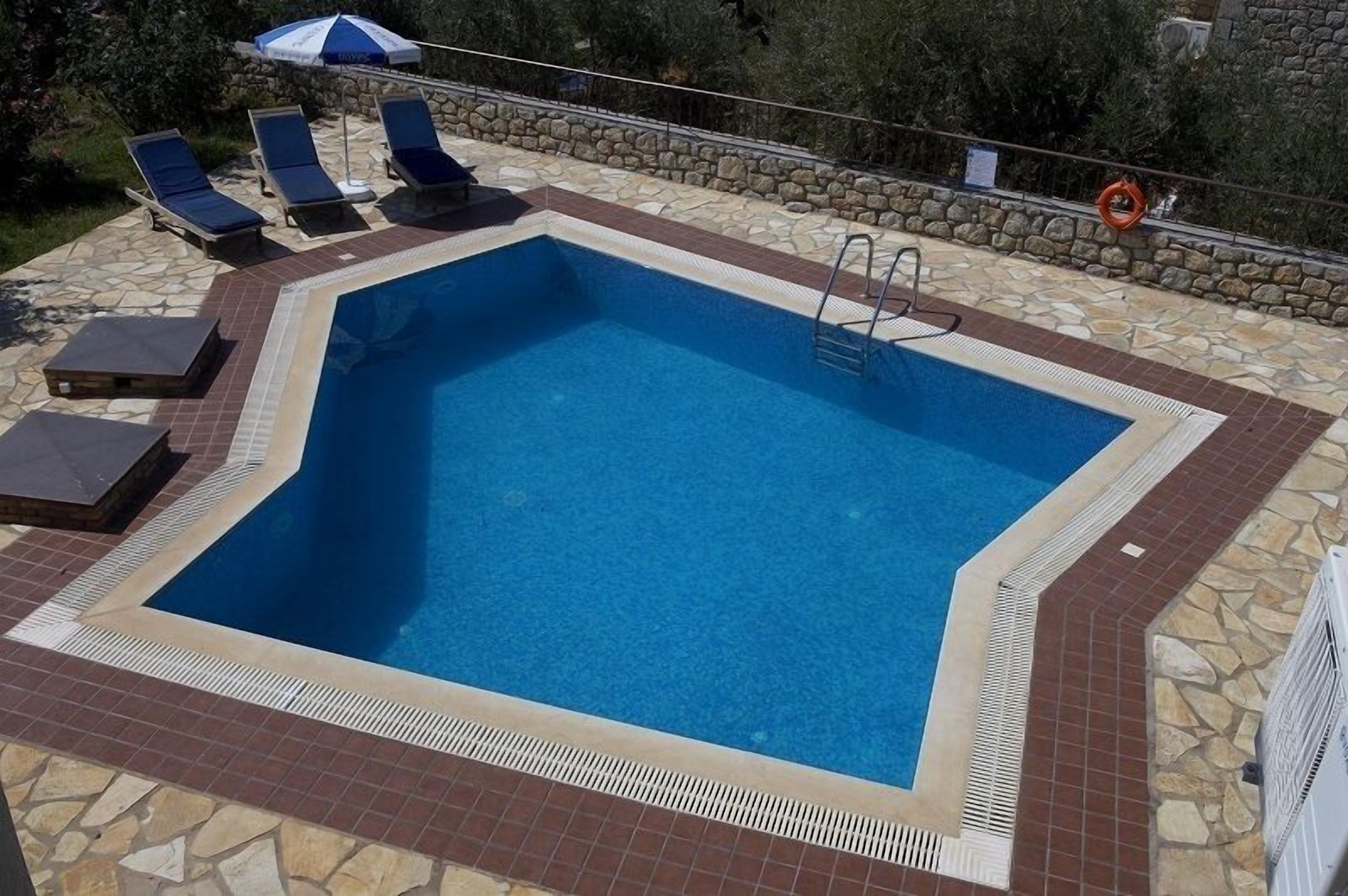 A rectangular swimming pool with a stone and tile border, surrounded by lounge chairs and a stone wall.