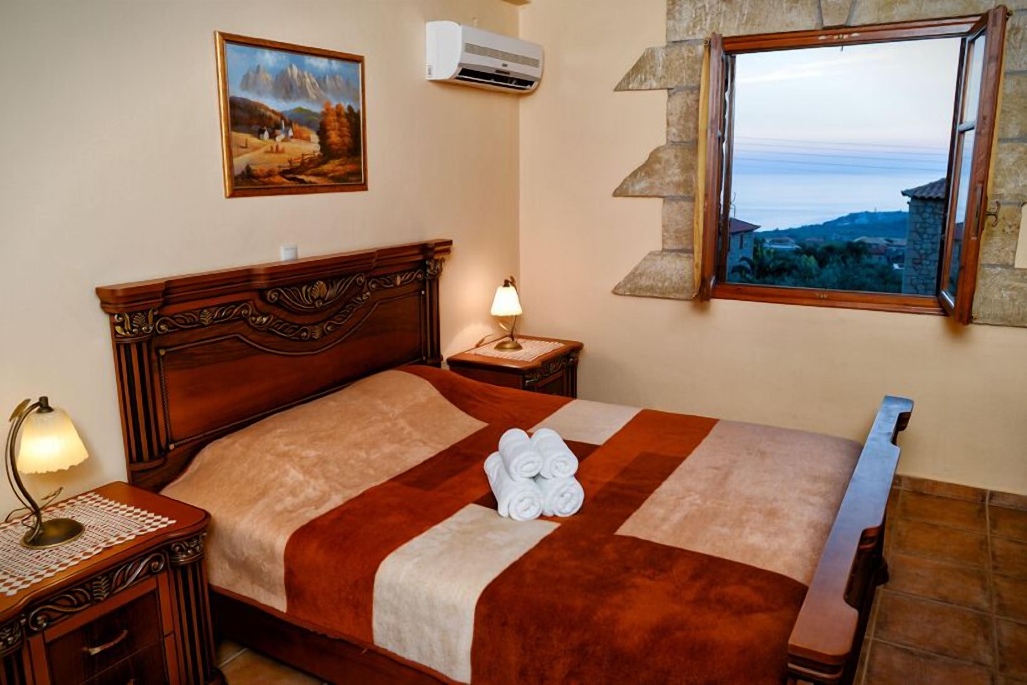A neatly made bed with a brown and beige quilt, a bedside lamp, a framed painting on the wall, and a window with a stone frame overlooking a landscape.
