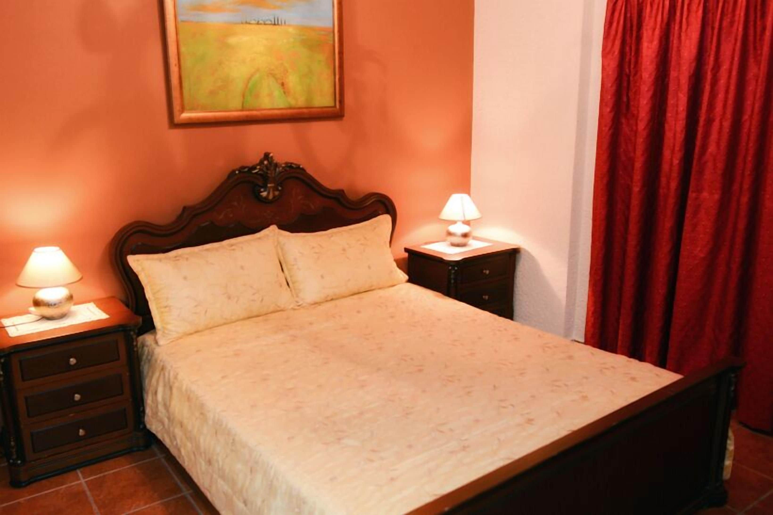 A bed with a wooden headboard, two bedside tables with lamps, a red curtain, and a painting on the wall.