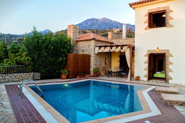 A swimming pool with a stone deck, a patio area with a table and chairs, and a mountain in the background. - Olympia Villas (West Mani)