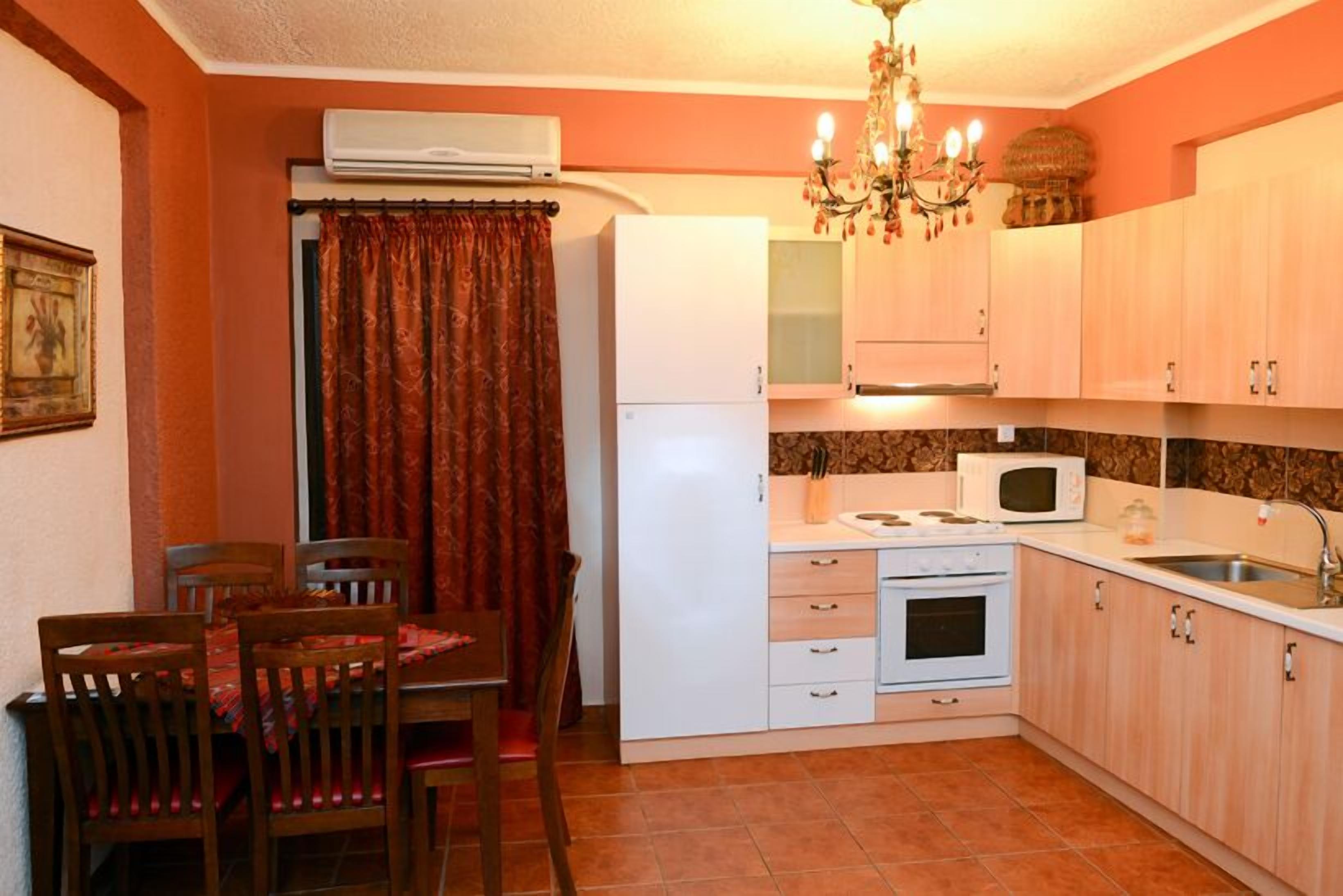 A compact kitchen with a refrigerator, microwave, stove, and sink. There is a dining table with chairs and a chandelier above.