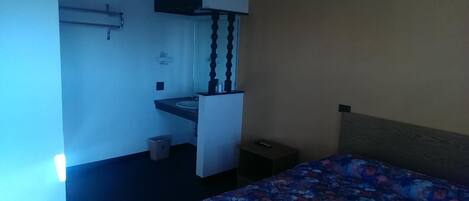 A small hotel room with a bed, a bedside table, a nightstand, a built-in desk with a chair, a small trash bin, and a wall-mounted shelf.