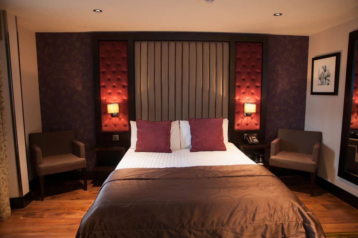 Hotellihuone, jossa on suuri sänky, kaksi nojatuolia, yöpöytä lampun kanssa ja seinälle asennettu puhelin.