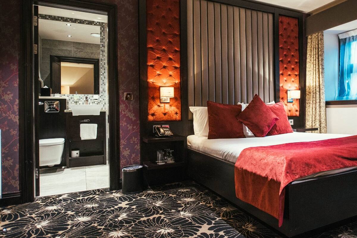 Moderni hotellihuone, jossa on suuri sänky, yöpöytä, peili ja näkymä kylpyhuoneeseen.