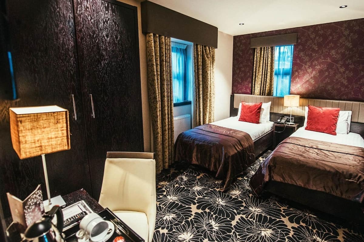 Hotellihuone, jossa on kaksi sänkyä, kirjoituspöytä lampun kanssa, tuoli ja kuviollinen matto.