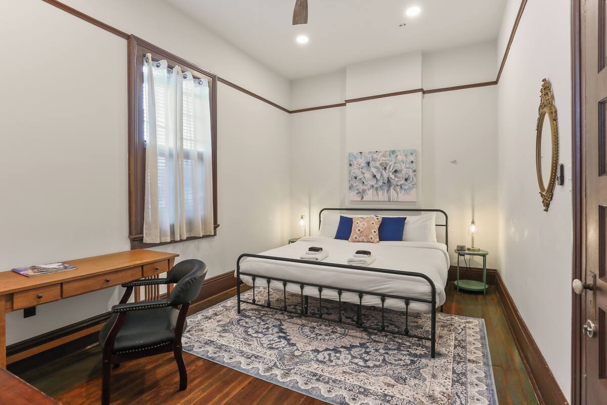 Un dormitorio con cama, escritorio, silla, alfombra estampada y un cuadro en la pared.