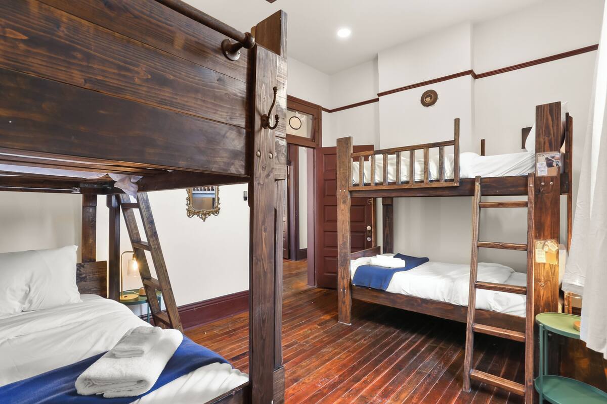 Una habitación con dos literas, piso de madera, una escalera, una pequeña mesa y una puerta que da a otra habitación.
