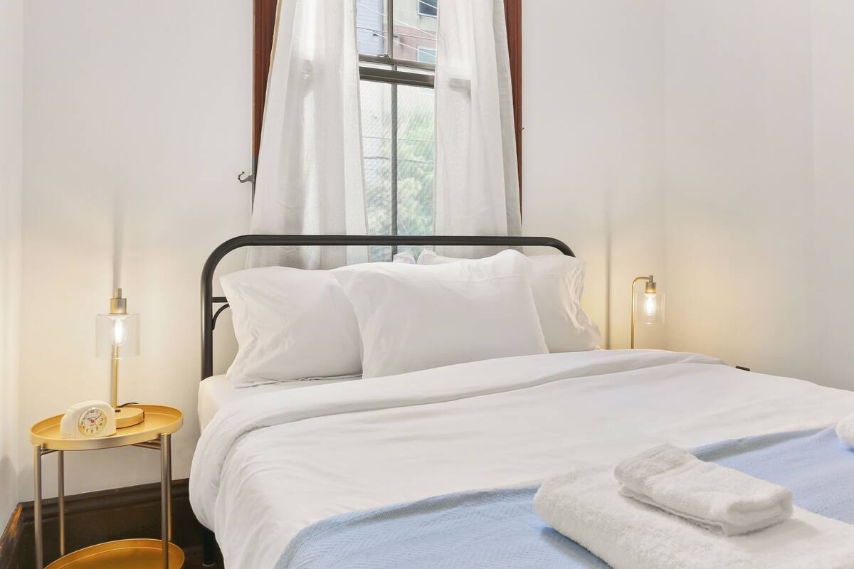 Una cama bien hecha con ropa blanca, una mesita de noche con lámpara y una ventana con cortinas traslúcidas.