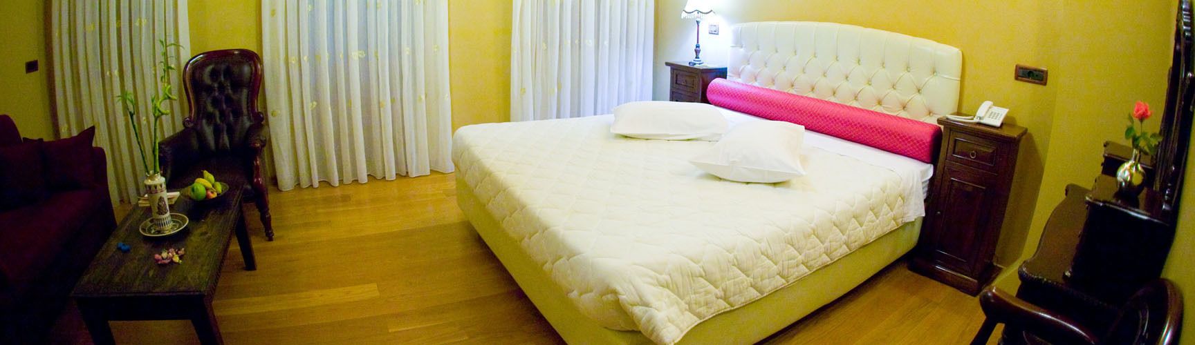 Una habitación de hotel con una cama grande, una mesita de noche de madera, un escritorio con un florero y una lámpara, y una silla.