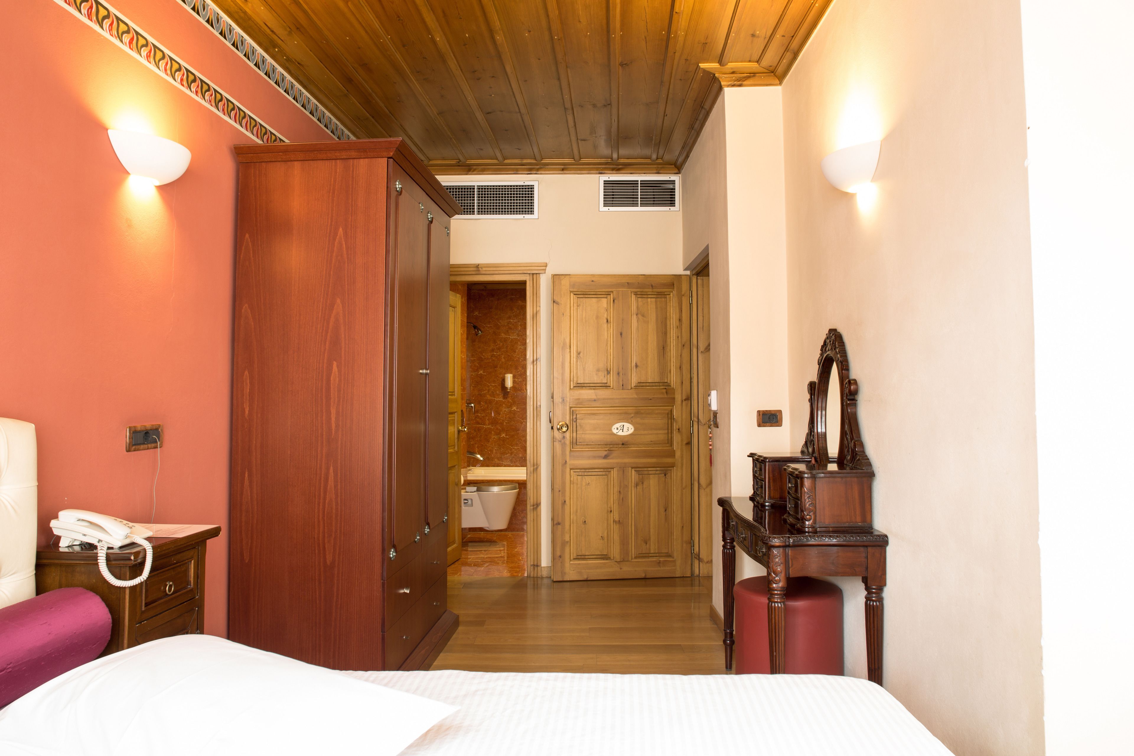A hotel room with a wooden wardrobe, a bedside telephone, a wooden dresser, and a mirror.
