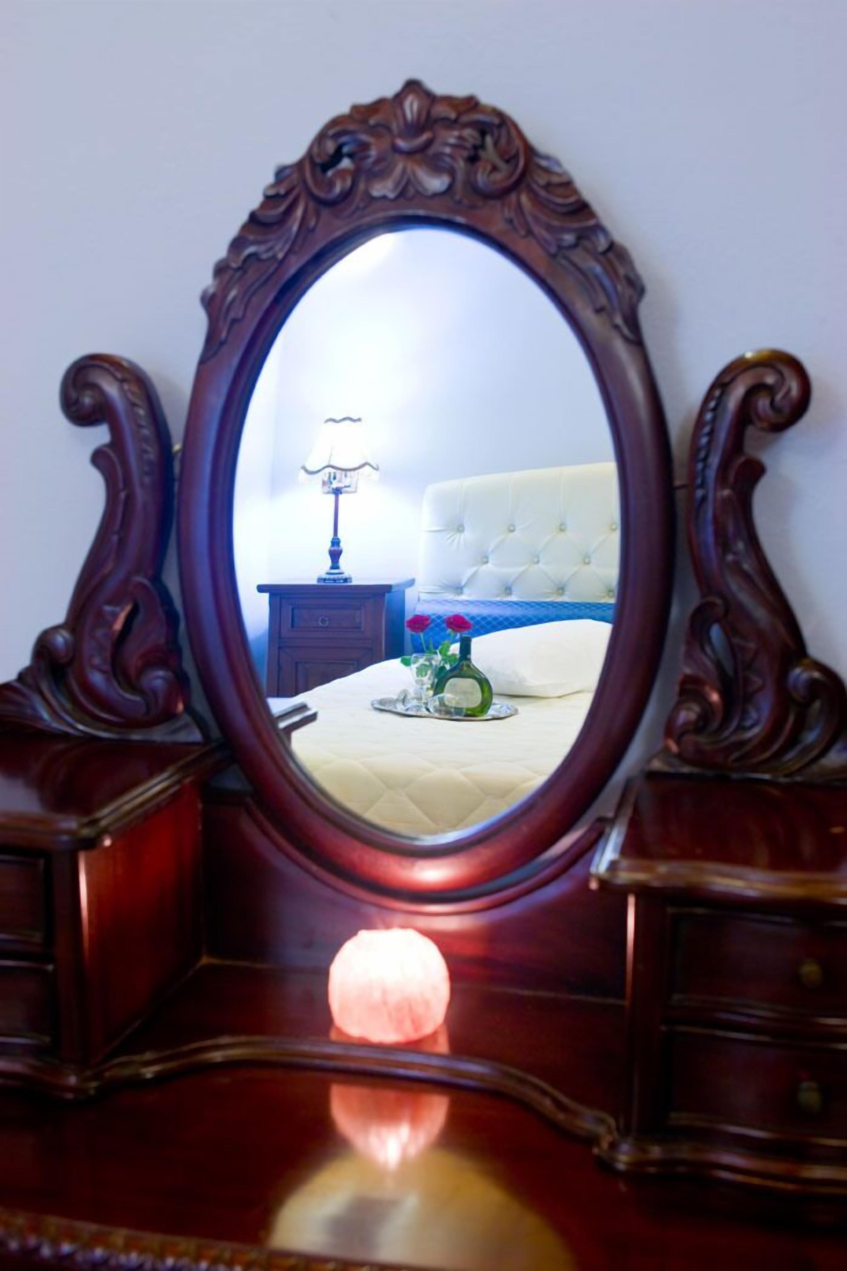 An ornate wooden-framed mirror reflecting a bedroom with a tufted headboard, a bedside table with a lamp, and a vase with flowers.