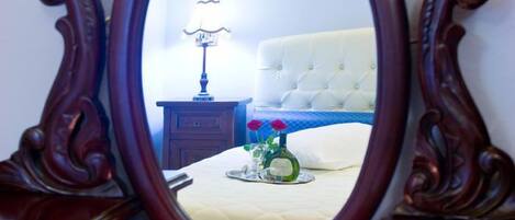 An ornate wooden-framed mirror reflecting a bedroom with a tufted headboard, a bedside table with a lamp, and a vase with flowers.