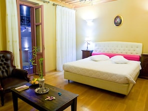 A bedroom with a bed, a nightstand, a wooden coffee table, a chair, and a window with curtains. - Kyveli Suites (Nafplio)