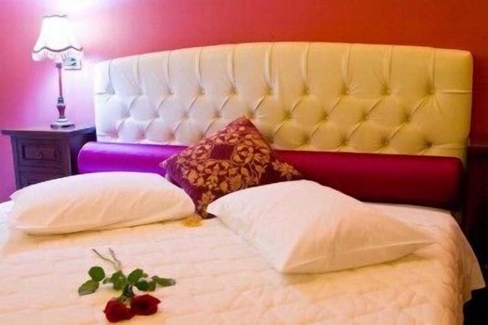 A bed with a tufted headboard, two white pillows, a red patterned pillow, and a single red rose on the bed.