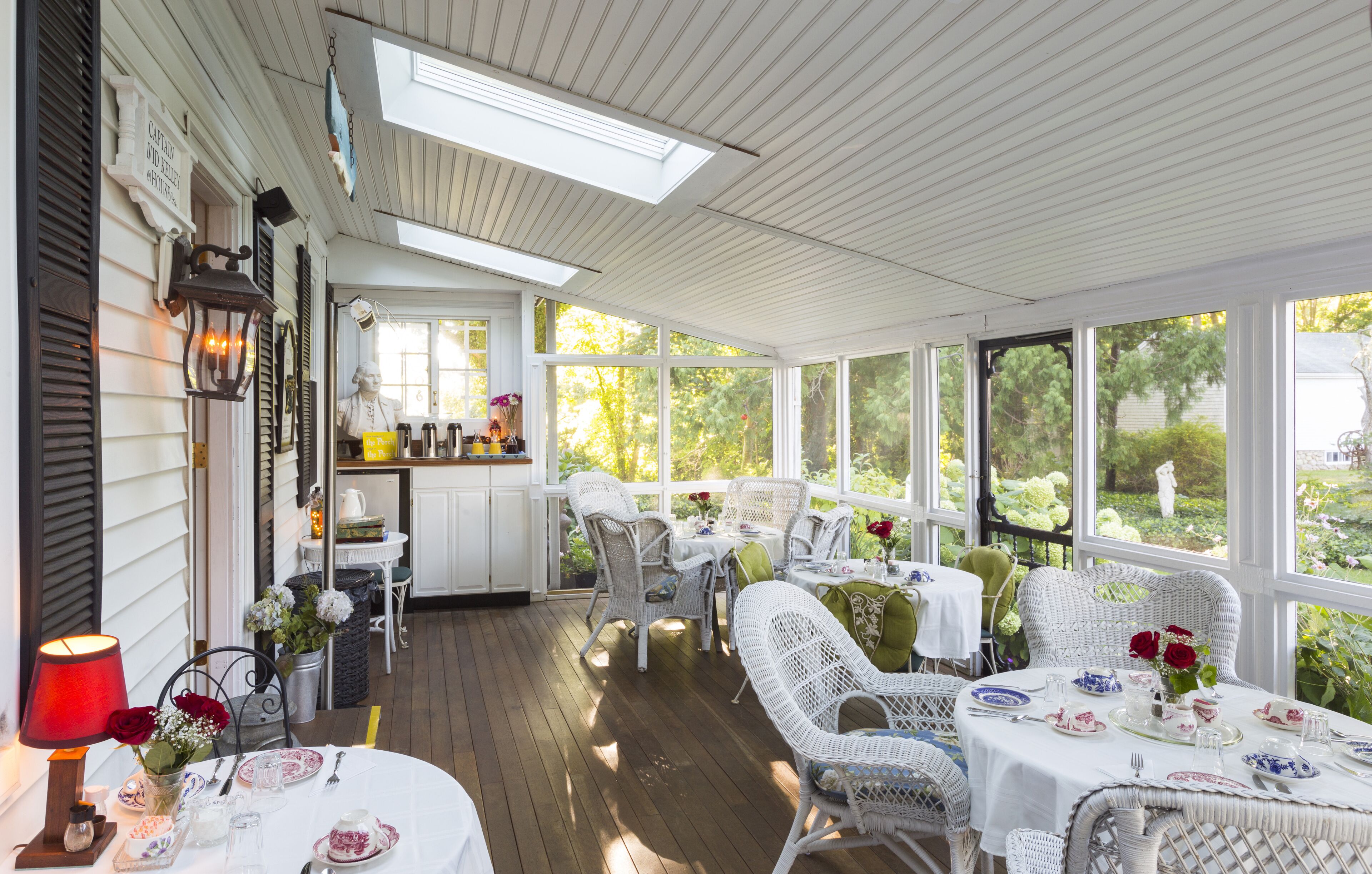 Un porche cubierto con muebles de mimbre blanco, una mesa de comedor con platos y flores, y vistas al follaje exterior.