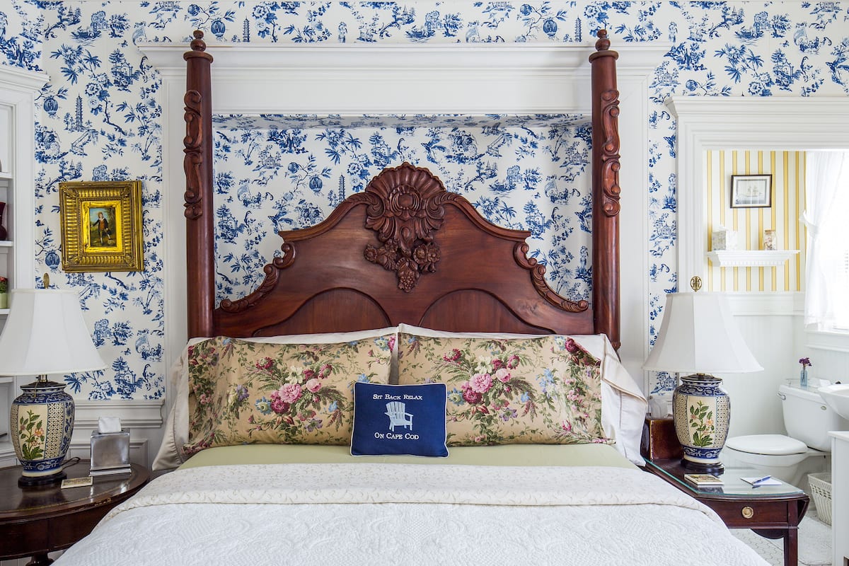 Une chambre à coucher avec un lit à baldaquin, un papier peint à motifs floraux et un coussin orné de l’inscription « St. Bede, détente sur Cape Cod ».