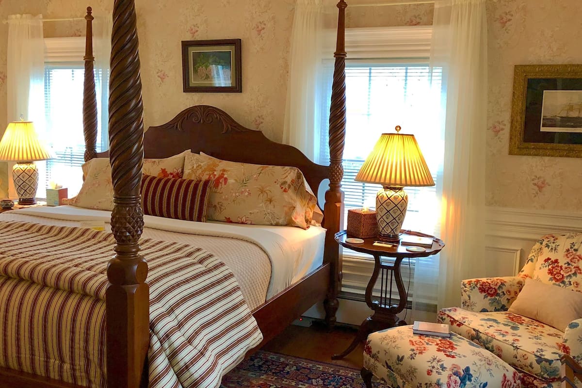 Un lit à baldaquin avec une couvre-lit à motifs floraux, un fauteuil assorti, une petite table ronde avec une lampe et une grande fenêtre avec des rideaux.