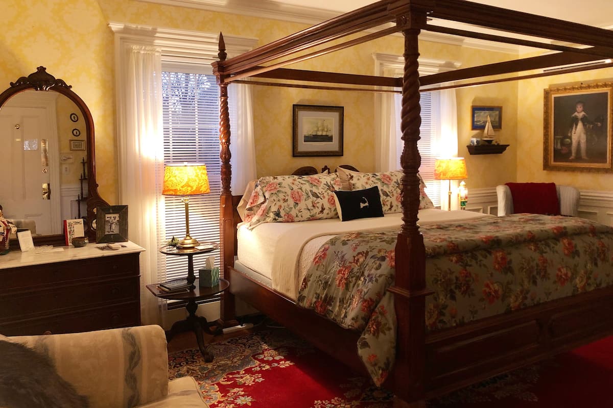 Un lit à baldaquin avec une literie à motifs floraux, une table de chevet en bois avec une lampe, une moquette rouge et un miroir au mur.
