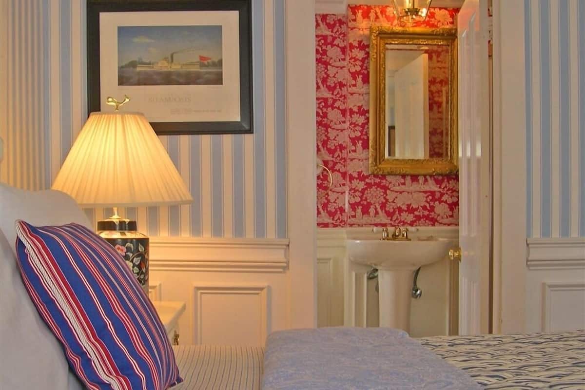 Une chambre d’hôtel avec un lit, une lampe de chevet, un tableau encadré au mur, une salle de bain avec un lavabo et un miroir, et un papier peint à rayures.