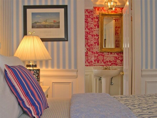 A hotel room with a bed, a bedside lamp, a framed picture on the wall, a bathroom with a sink and mirror, and striped wallpaper. - Captain David Kelley House Bed & Breakfast (Centerville)