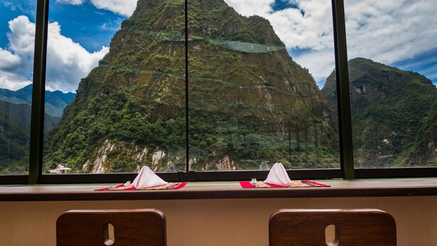 Un comedor con sillas de madera y una mesa, una ventana con vista a la montaña, y un portavasos con un pañuelo rojo.