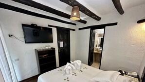 Habitación de hotel con cama, televisor montado en la pared, repisas con artículos de aseo y una puerta que da al baño.