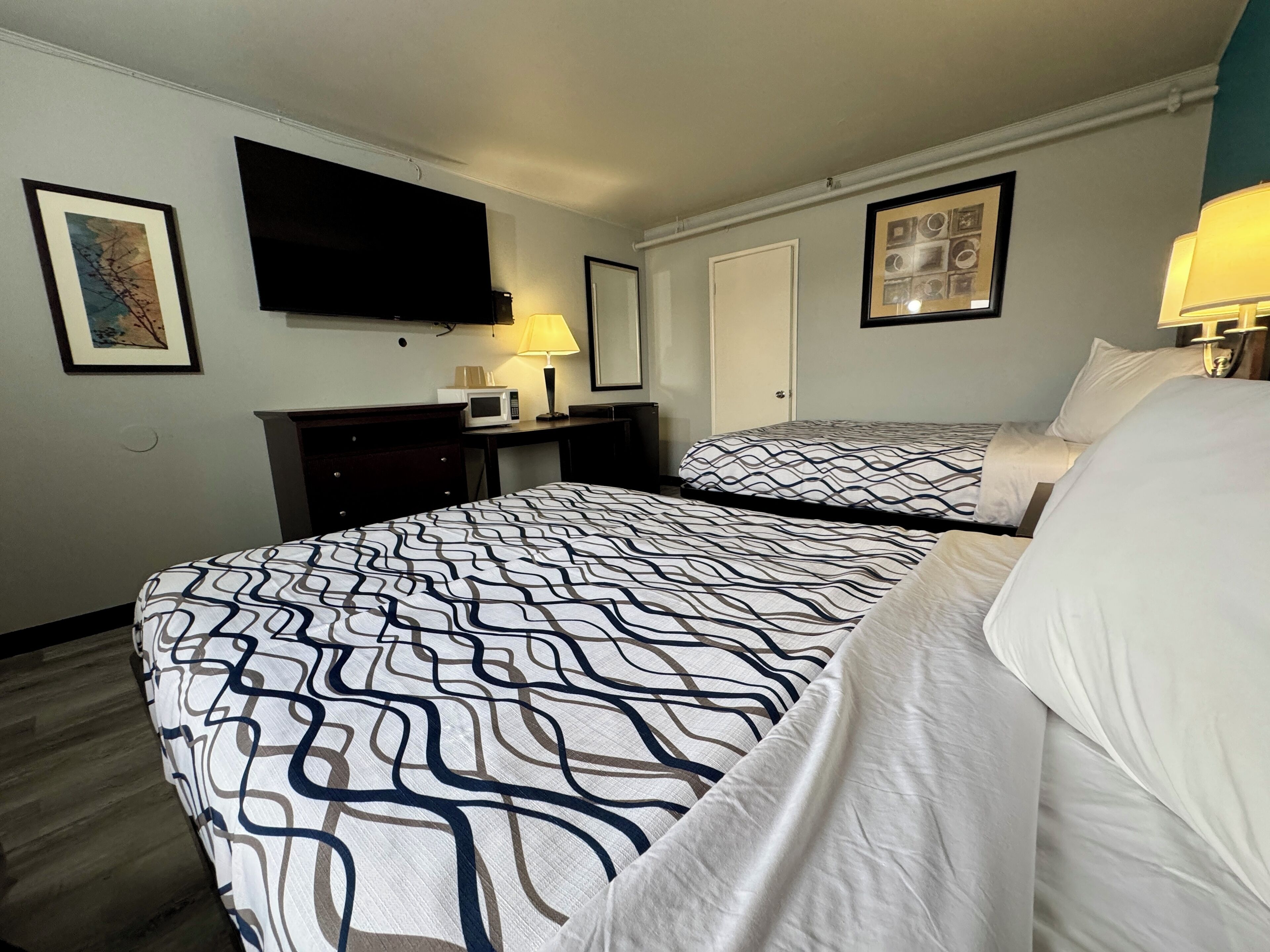 A hotel room with two beds, a flat-screen TV mounted on the wall, a desk with a lamp, and framed artwork on the walls.
