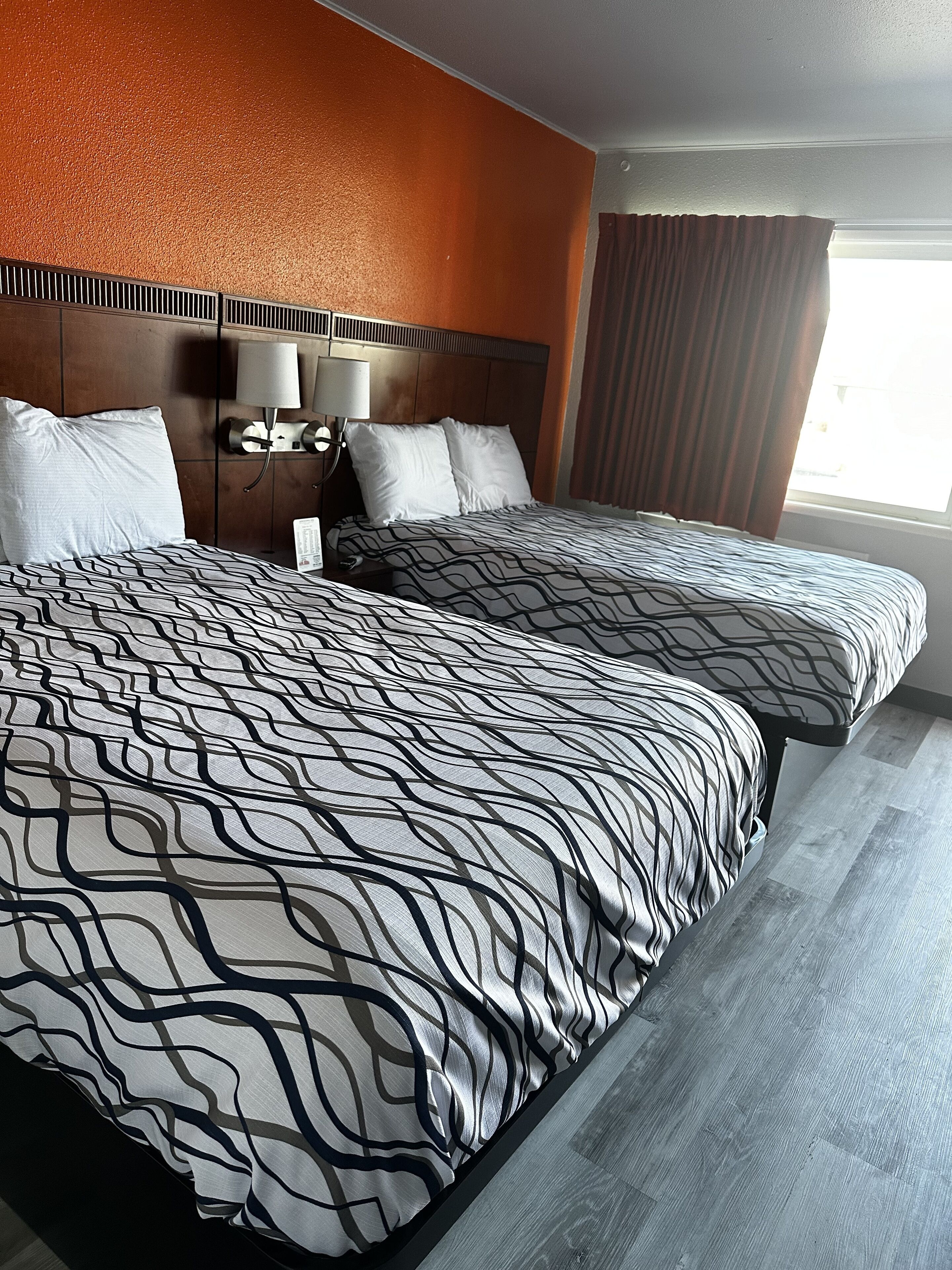 A hotel room with two beds, a headboard, a nightstand, a lamp, and a window with curtains.