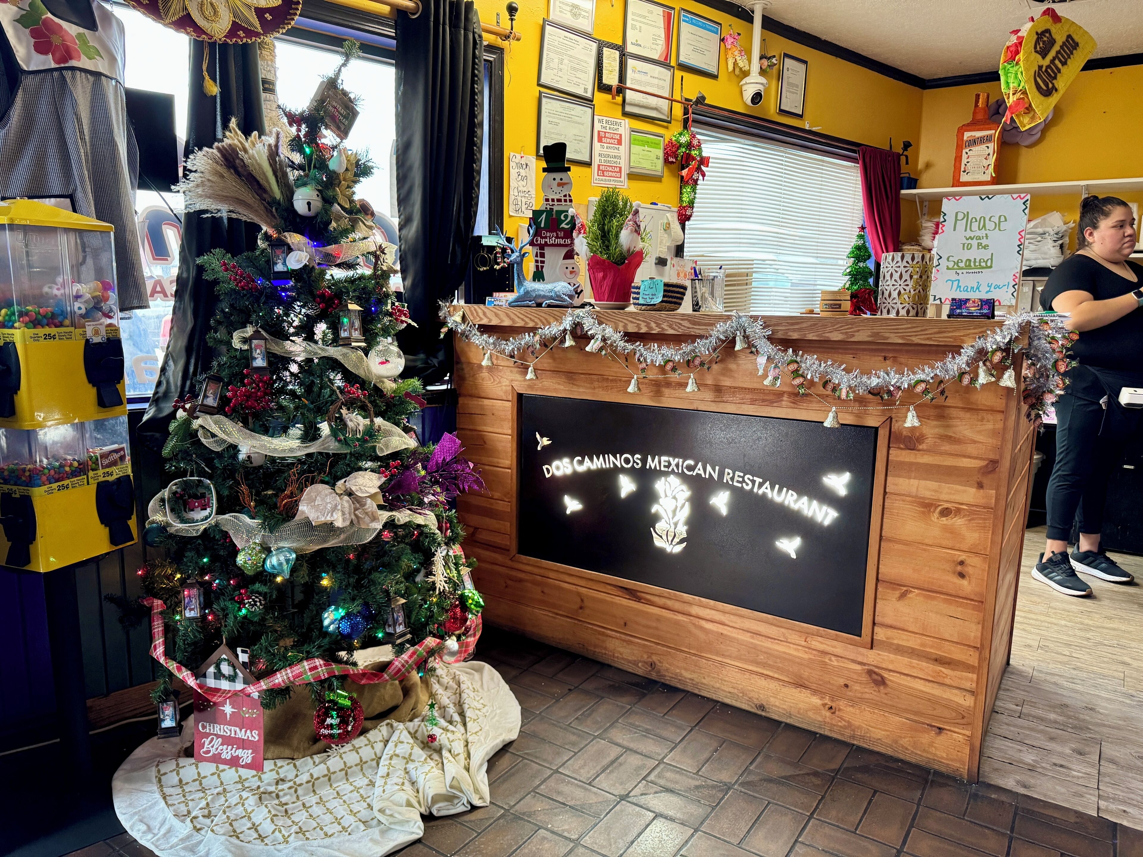 Interior de un restaurante con un árbol de Navidad, una barra de madera con el nombre 'DOS CAMINOS MEXICAN RESTAURANT', y un letrero que indica un 12% de descuento el día de Navidad.