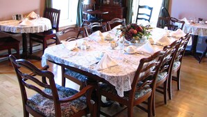 Ein Essbereich mit runden Tischen für vier Personen, Holzstühlen mit gepolsterten Sitzen, Spitze-Tischdecken und einer Blumenarrangements als Mittelpunkt.