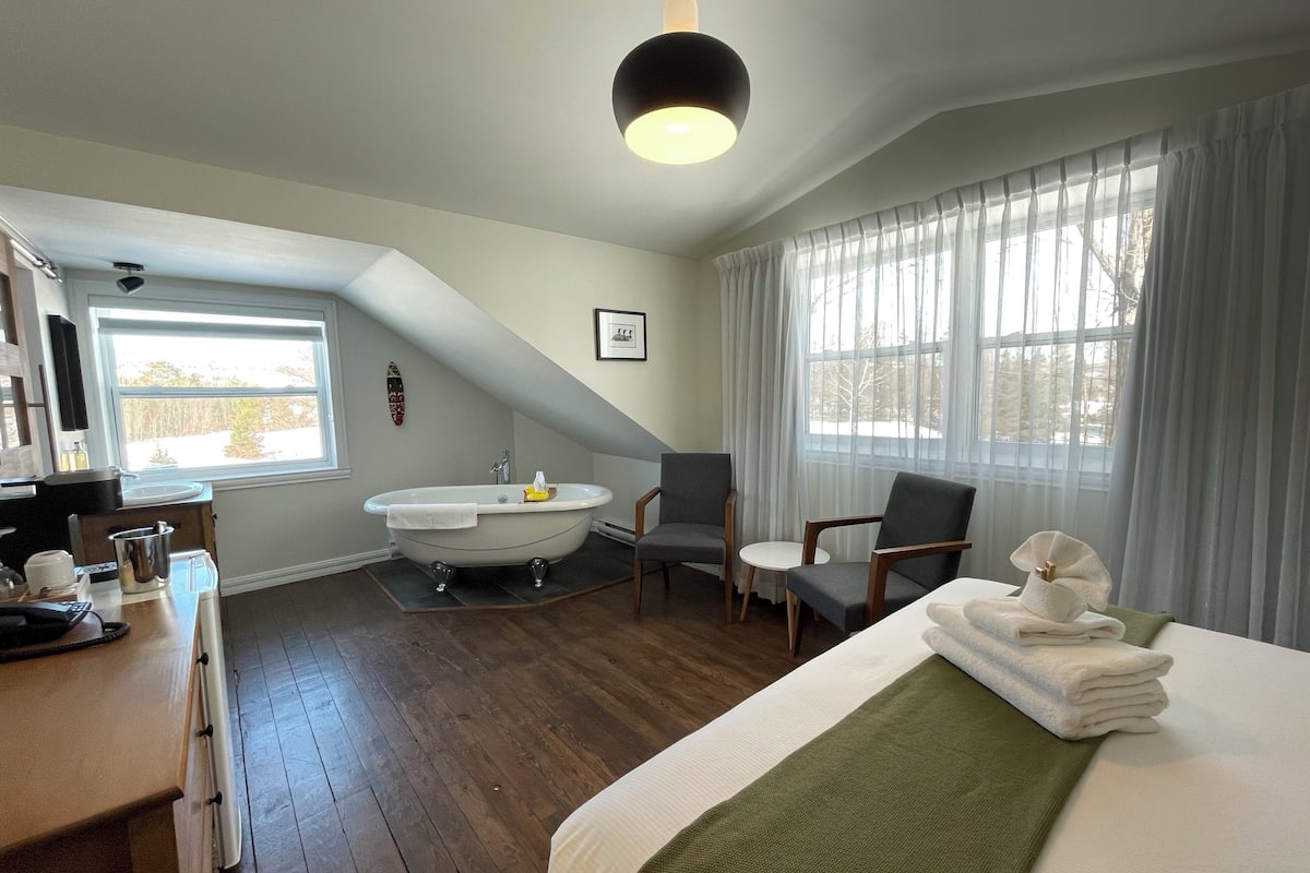 A bedroom with a large bed, a sitting area with a chair, a bathtub, and a window with curtains.