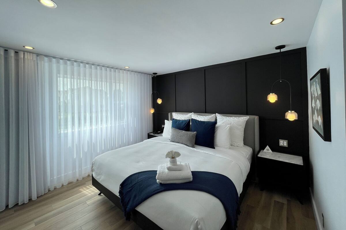 A modern hotel room with a large bed, a nightstand, a wall-mounted lamp, and a window with sheer curtains.