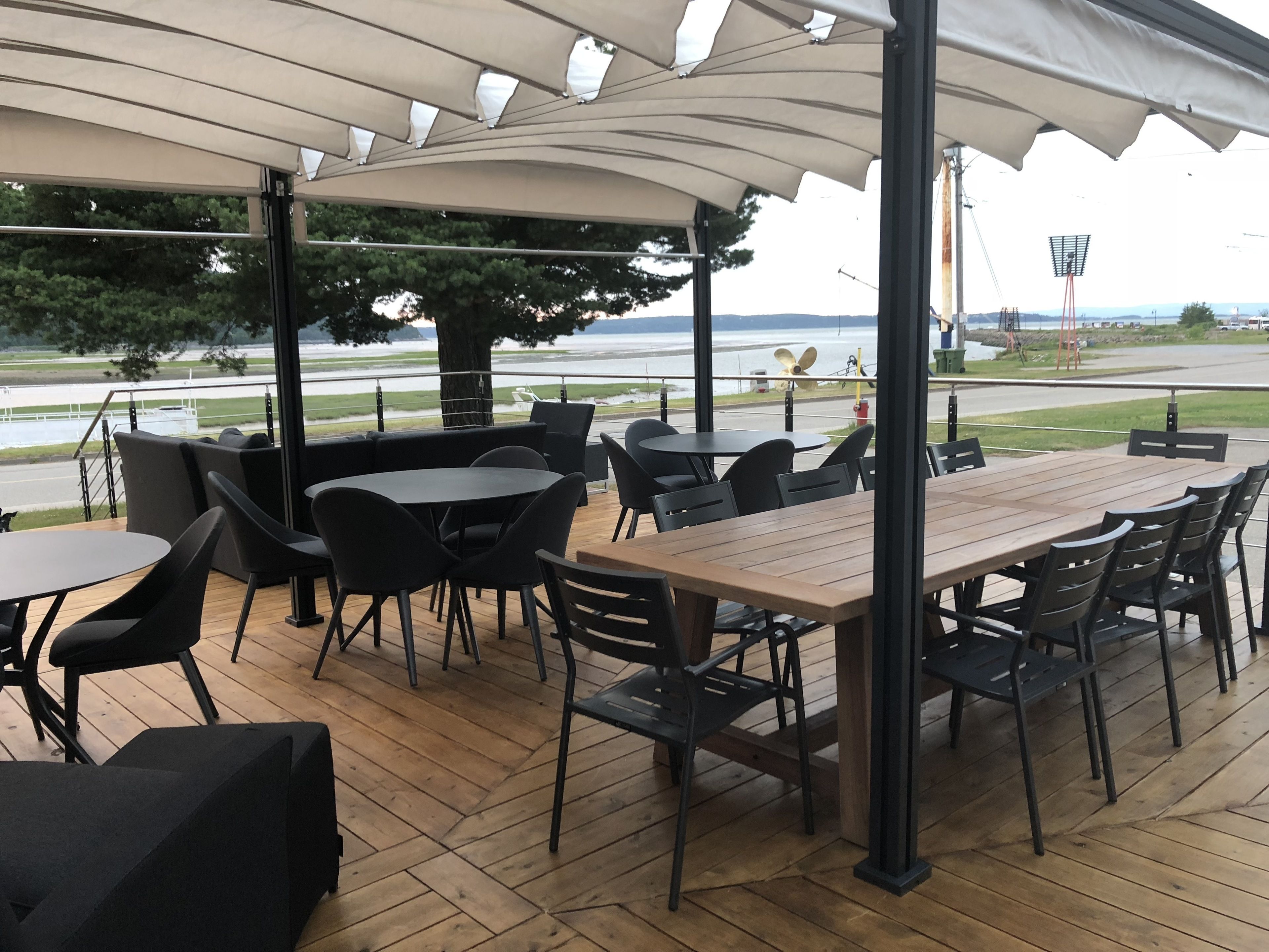 An outdoor patio with wooden tables and black chairs, a large tree, and a view of a body of water and distant landscape.