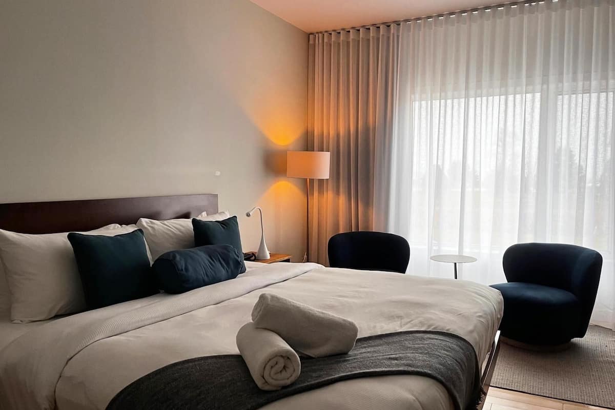 A hotel room with a bed, a nightstand with a lamp, two chairs, and a view of the outdoors through sheer curtains.