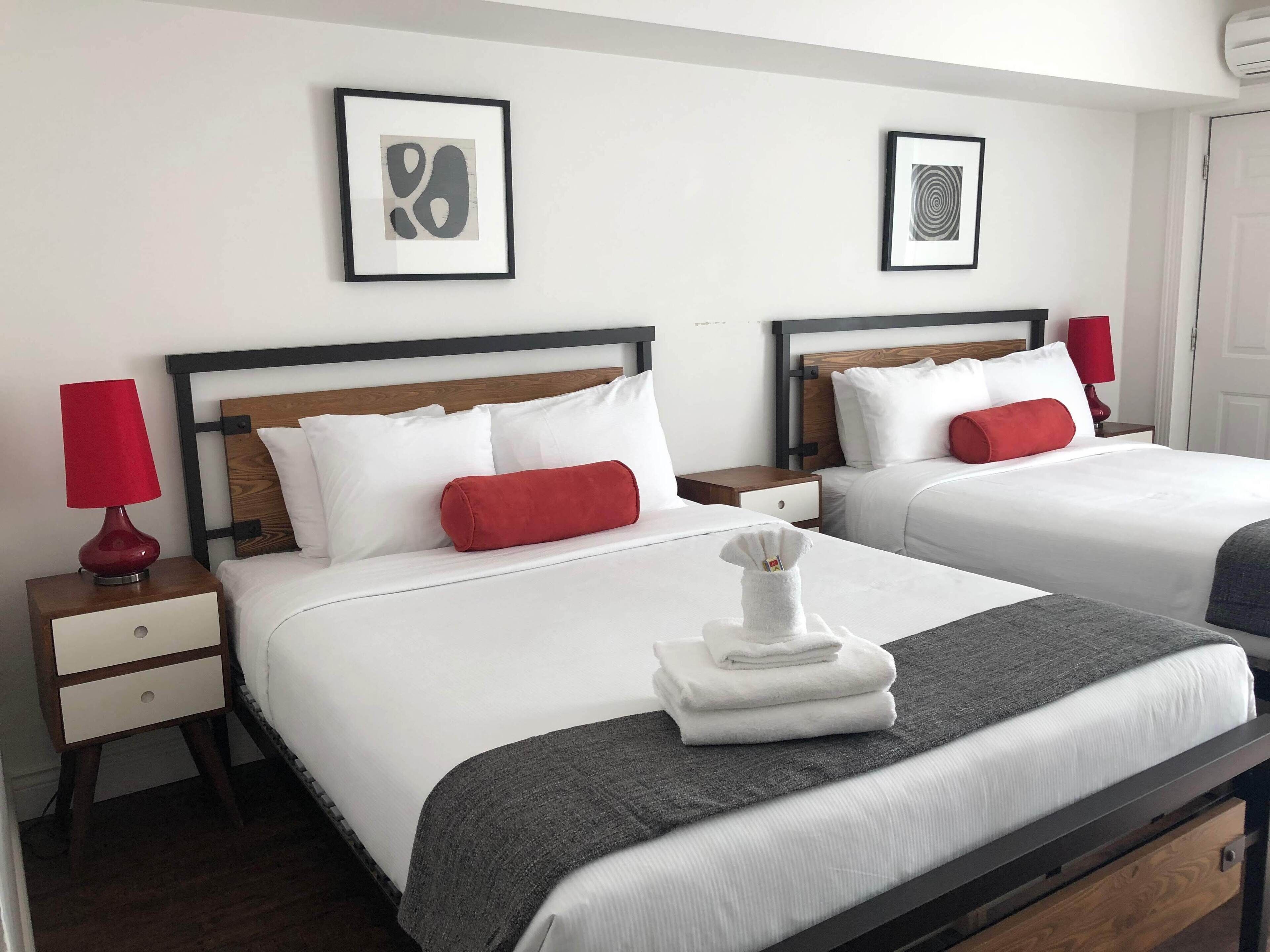 A hotel room with two beds, white bedding, red pillows, a grey throw blanket, a red lamp, a wooden nightstand, and a framed abstract artwork on the wall.