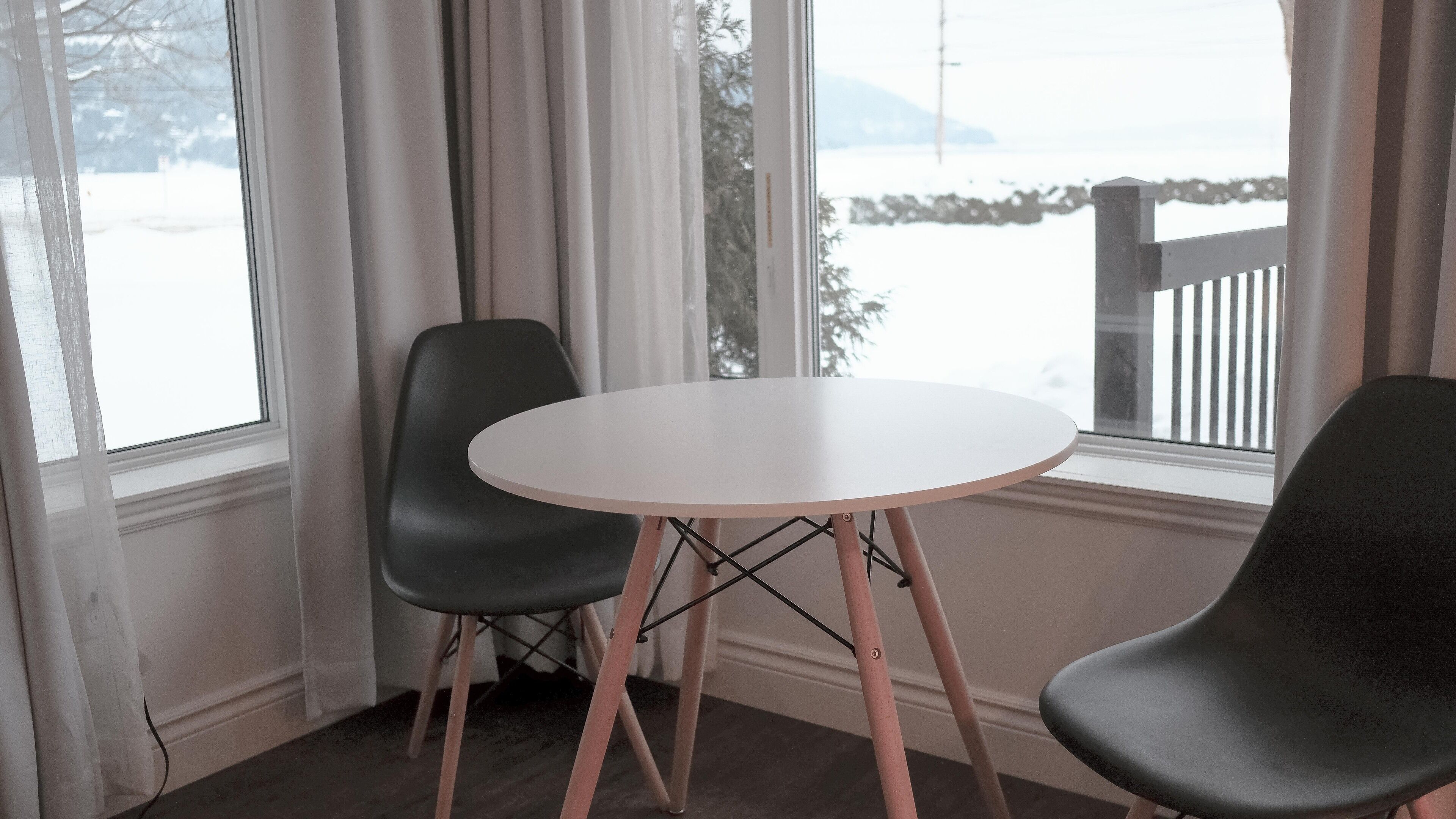 A room with a round table and two chairs, large windows with curtains, and a view of a snowy landscape.