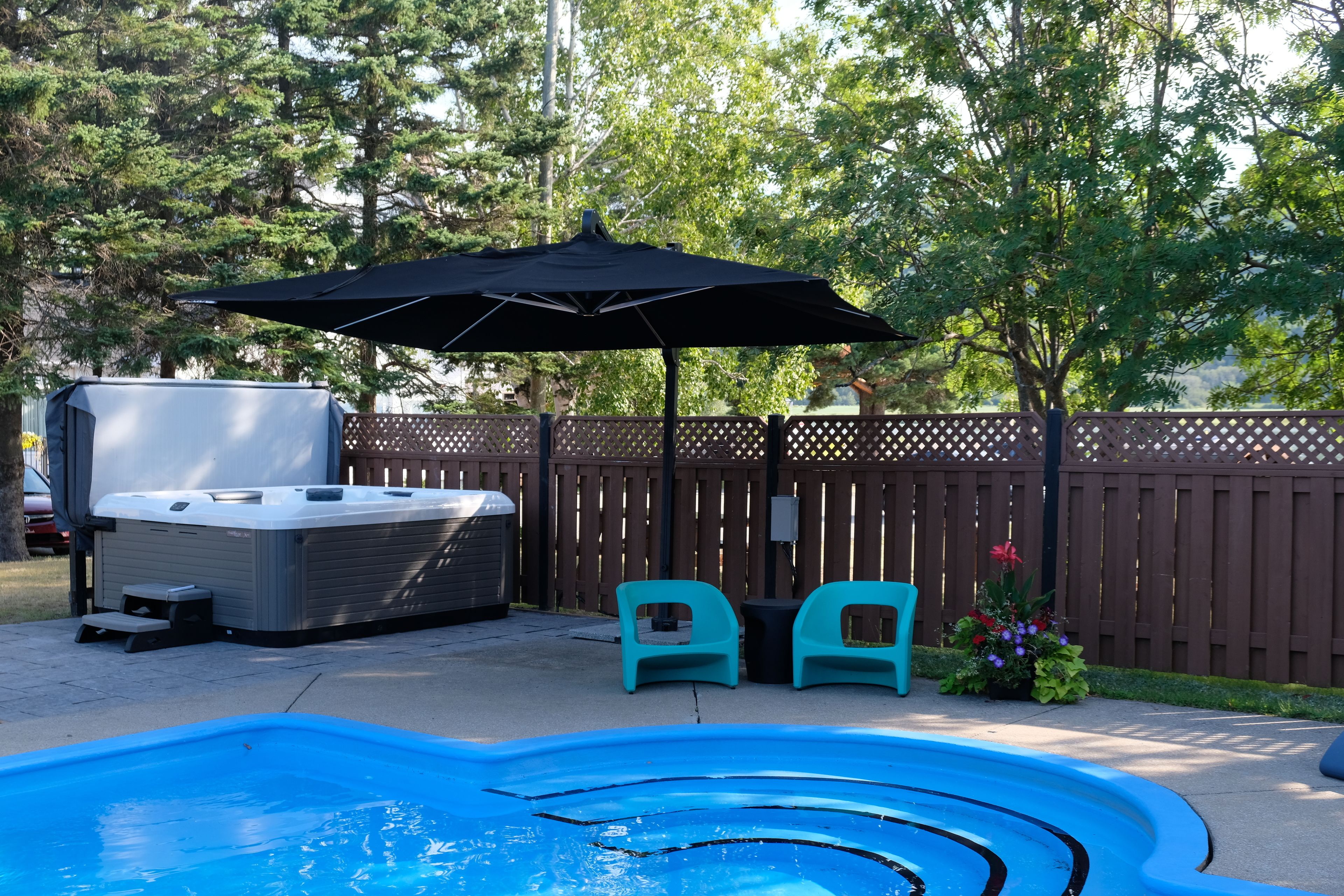 Ein Garten mit Schwimmbad, Whirlpool und zwei türkisblauen Stühlen unter einem schwarzen Sonnenschirm.
