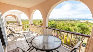 A balcony with a glass table and chairs, offering a view of a lush landscape and distant mountains.