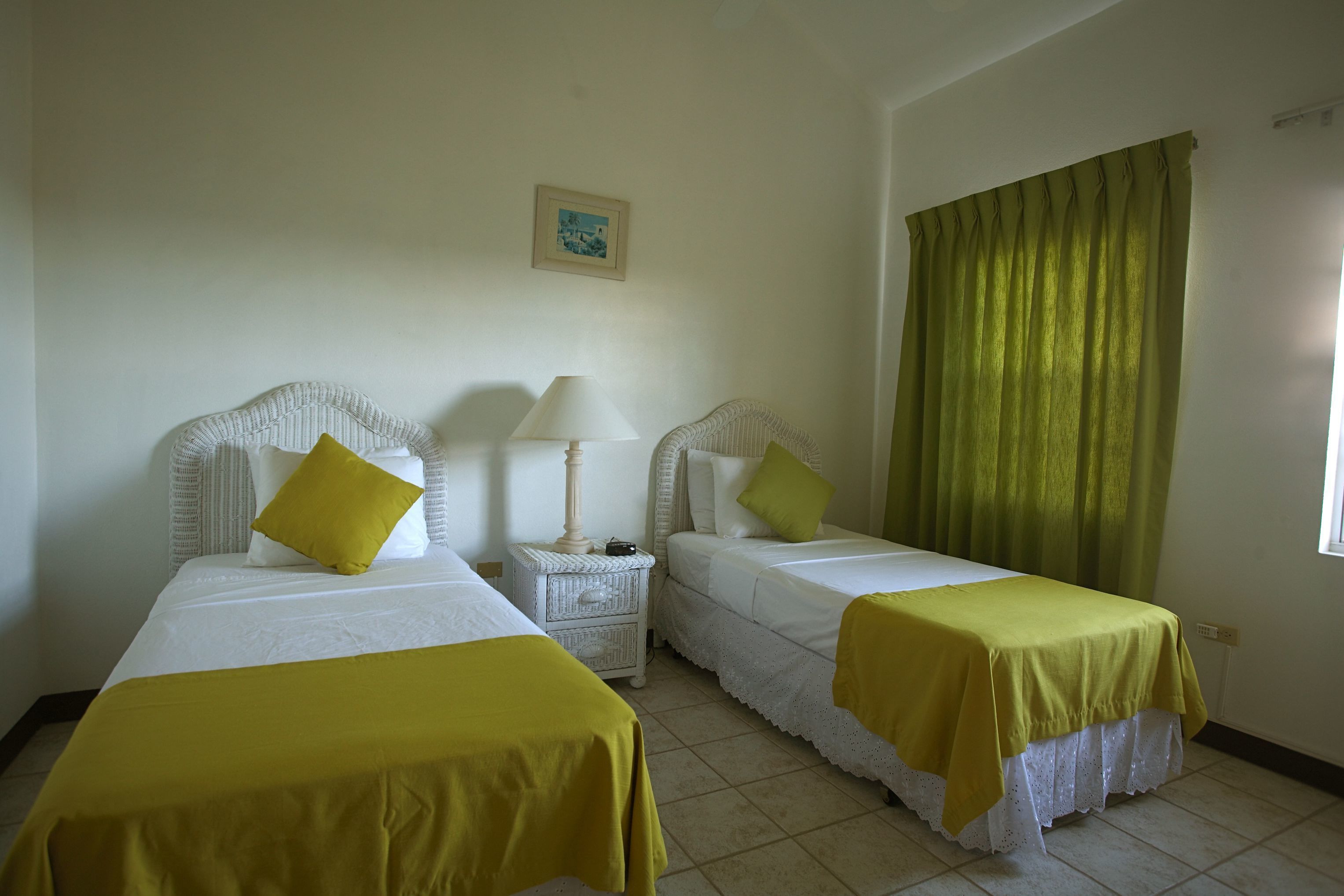 Habitación de hotel con dos camas, cada una con cabecera blanca y colcha amarilla. Hay una mesita de noche pequeña con una lámpara y un cuadro en la pared encima.