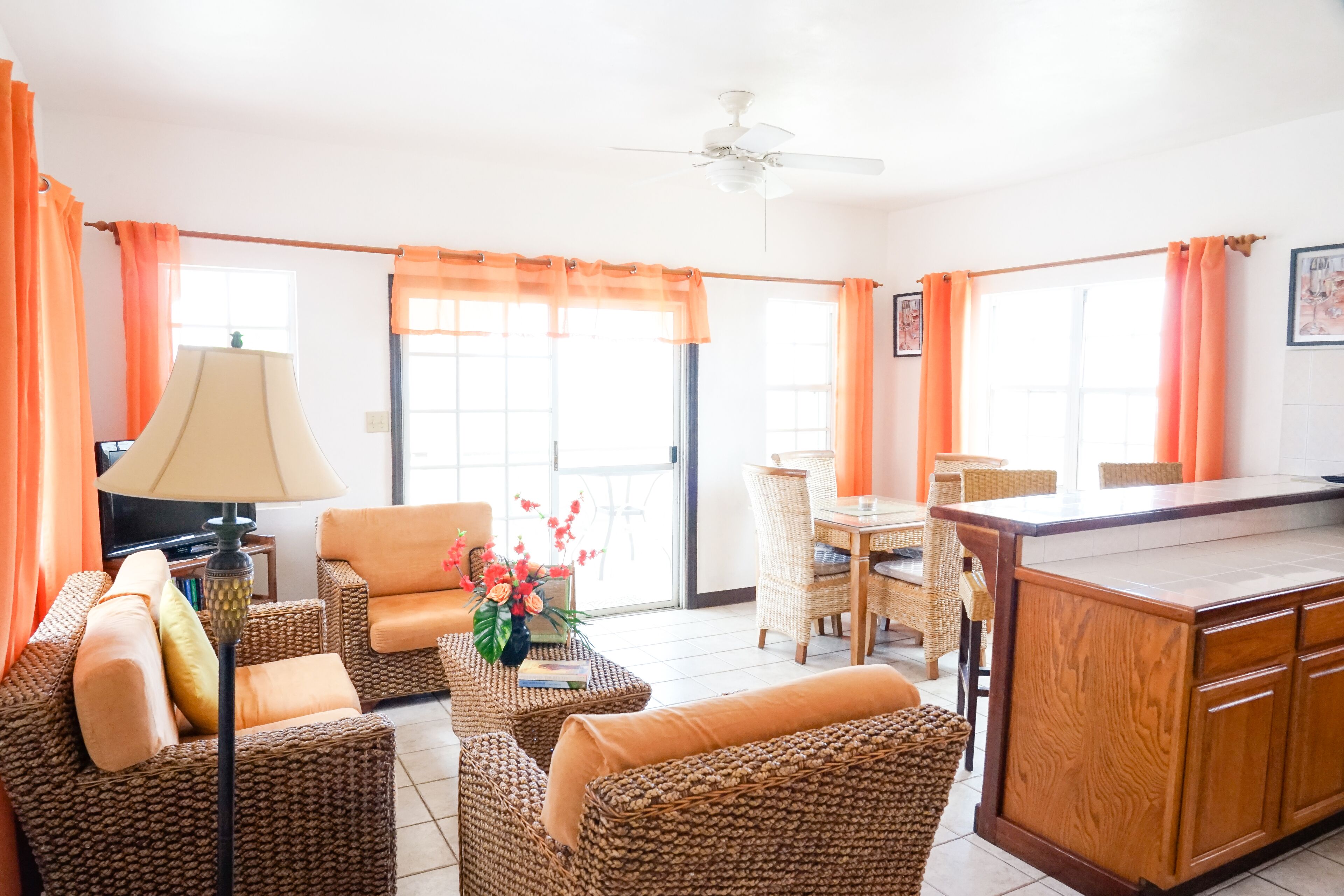 A bright living room with wicker furniture, a dining area, and a kitchen with a wooden island.
