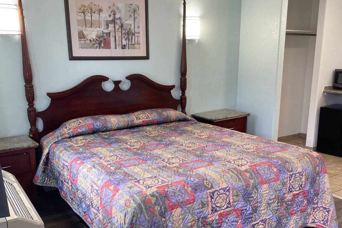 A bedroom with a wooden four-poster bed, a colorful quilt, two bedside tables, a framed picture on the wall, and a door leading to another room.