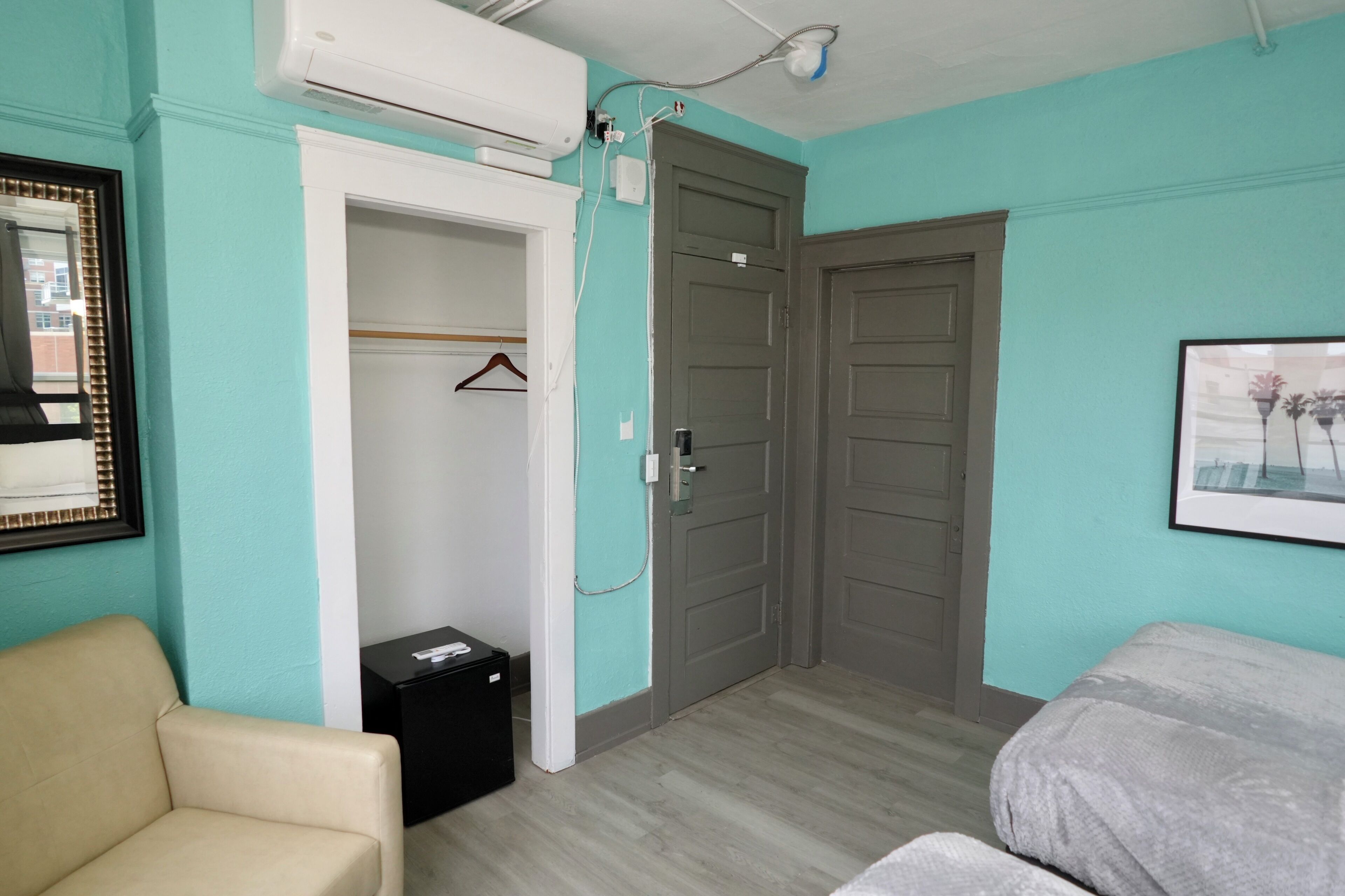 A room with teal walls, a beige chair, a bed, a wardrobe, and a framed picture on the wall.