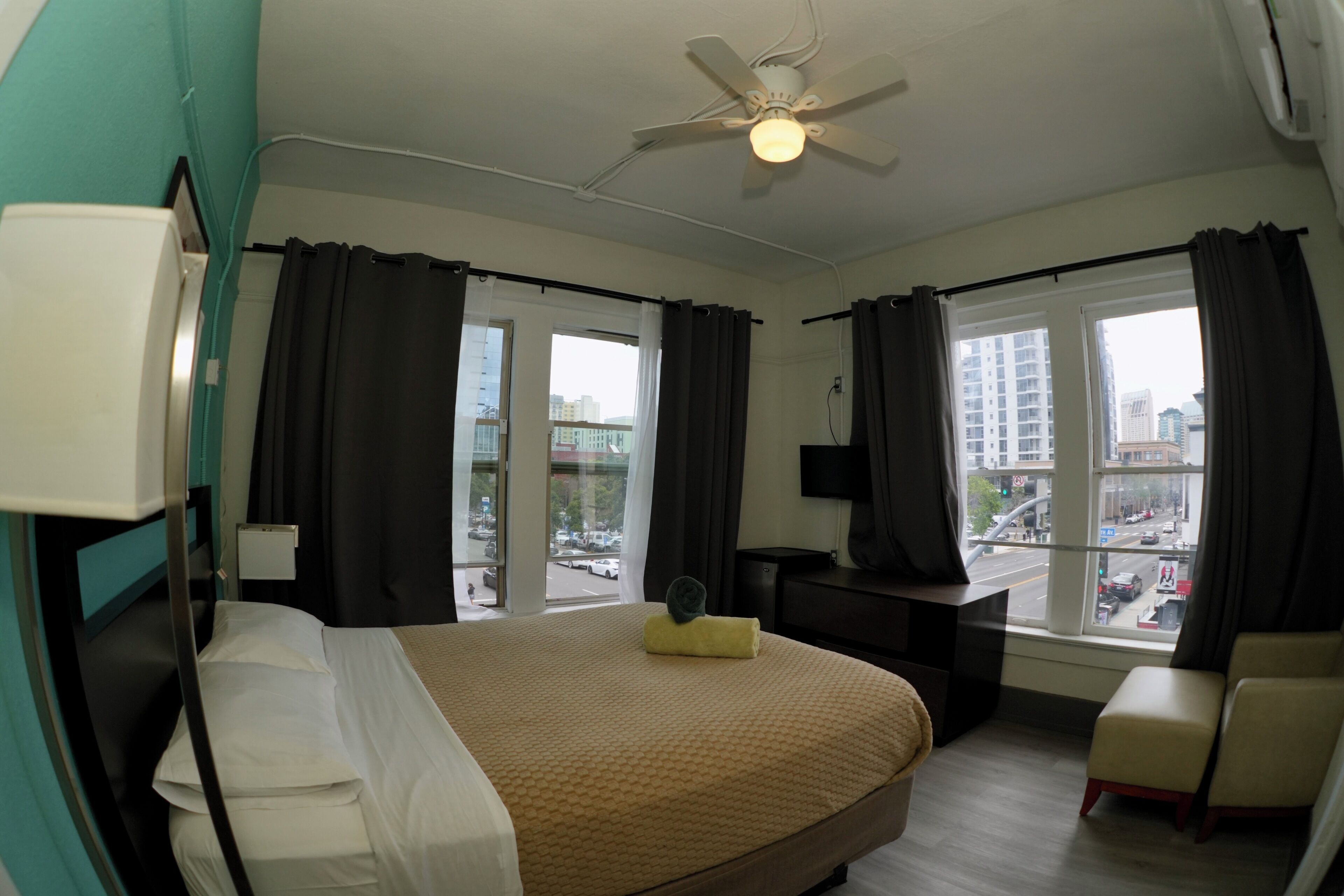 A hotel room with a bed, a desk, a chair, a ceiling fan, and large windows with curtains.