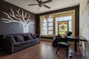A room with a dark brown sofa, a dining table with chairs, a ceiling fan, a stained glass window, and a wall mural.