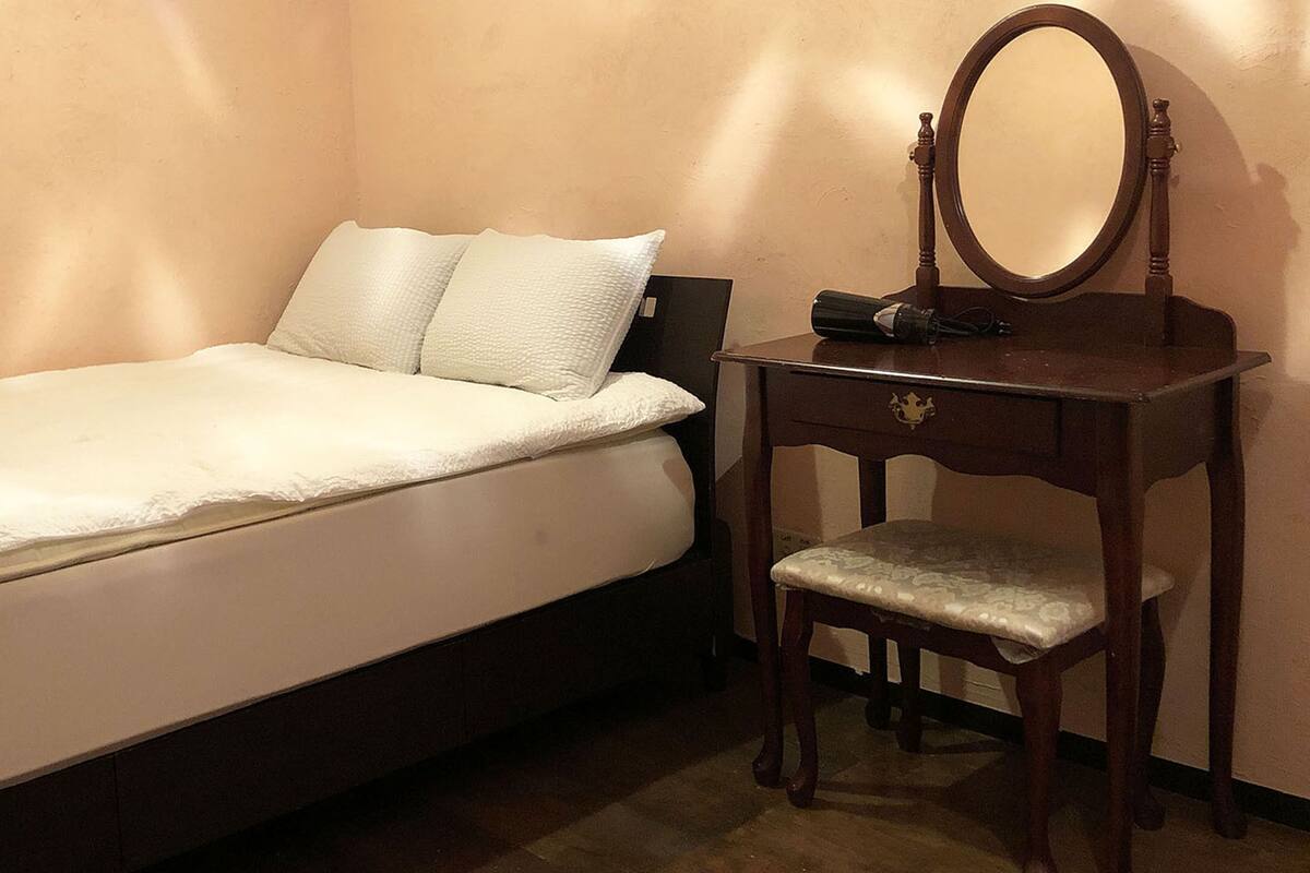 Una cama individual con un edredón blanco y almohadas, una mesita de noche de madera con espejo, y una silla con cojín.