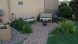 A paved garden area with two benches, a potted plant with yellow flowers, and a variety of greenery. - Brentwood Inn & Suites (Rochester)