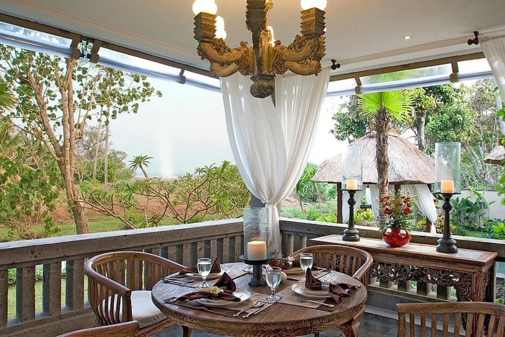 An outdoor dining area with a round table, chairs, a chandelier, and a view of the surrounding greenery.